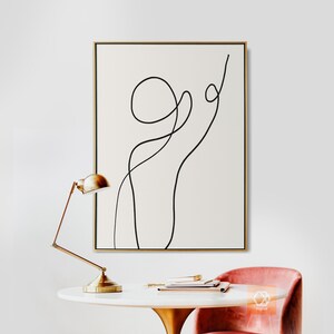 May include: A black and white abstract line art print in a gold frame hanging on a white wall. A gold desk lamp and a pink chair are in the foreground.