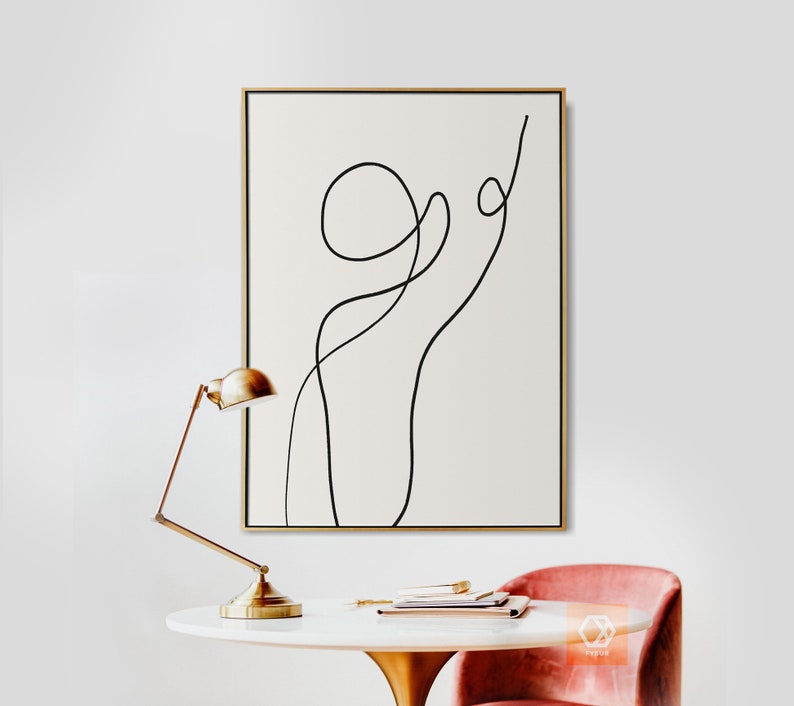 May include: A black and white abstract line art print in a gold frame hanging on a white wall. A gold desk lamp and a pink chair are in the foreground.