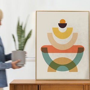 May include: Abstract art print with a gold frame. The artwork features a geometric design with orange, yellow, green, and brown shapes on a cream background. A person is holding a potted plant near the artwork.