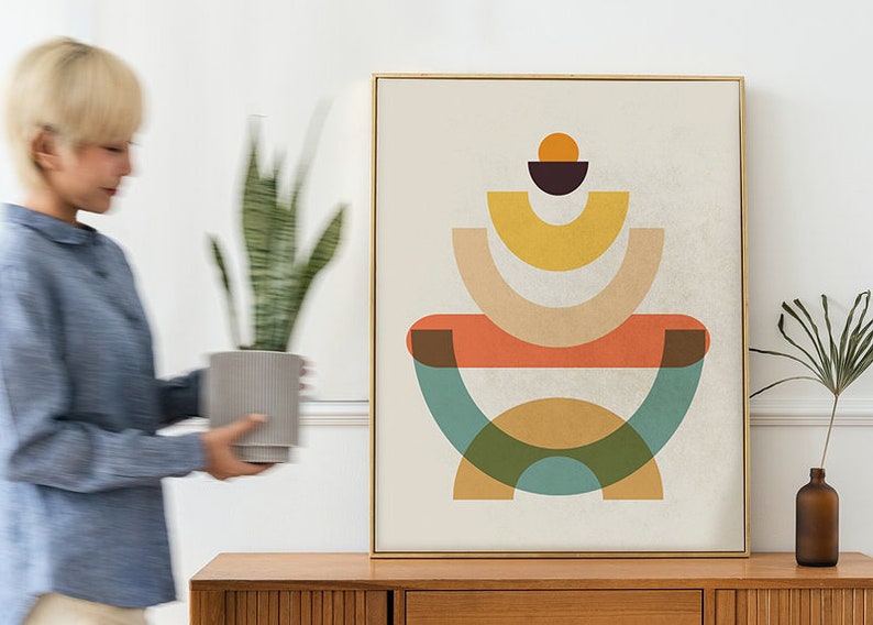 May include: Abstract art print with a gold frame. The artwork features a geometric design with orange, yellow, green, and brown shapes on a cream background. A person is holding a potted plant near the artwork.