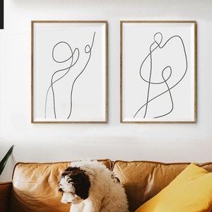May include: Two minimalist line art prints in black and white, framed in natural wood, hanging on a white wall. The prints feature abstract line drawings of figures.