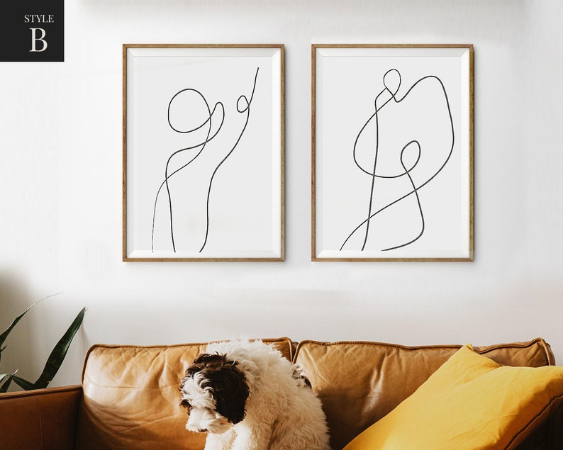 May include: Two minimalist line art prints in black and white, framed in natural wood, hanging on a white wall. The prints feature abstract line drawings of figures.