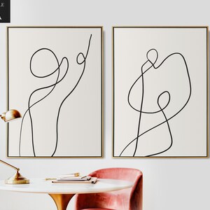 May include: Two minimalist line art prints in black and white on a white background. The prints are framed in gold and feature abstract line drawings.