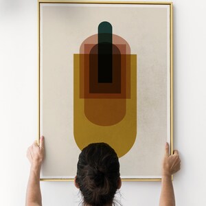 May include: A person hangs a framed abstract art print on a white wall. The print features a geometric design with overlapping shapes in shades of brown, yellow, red, and green.