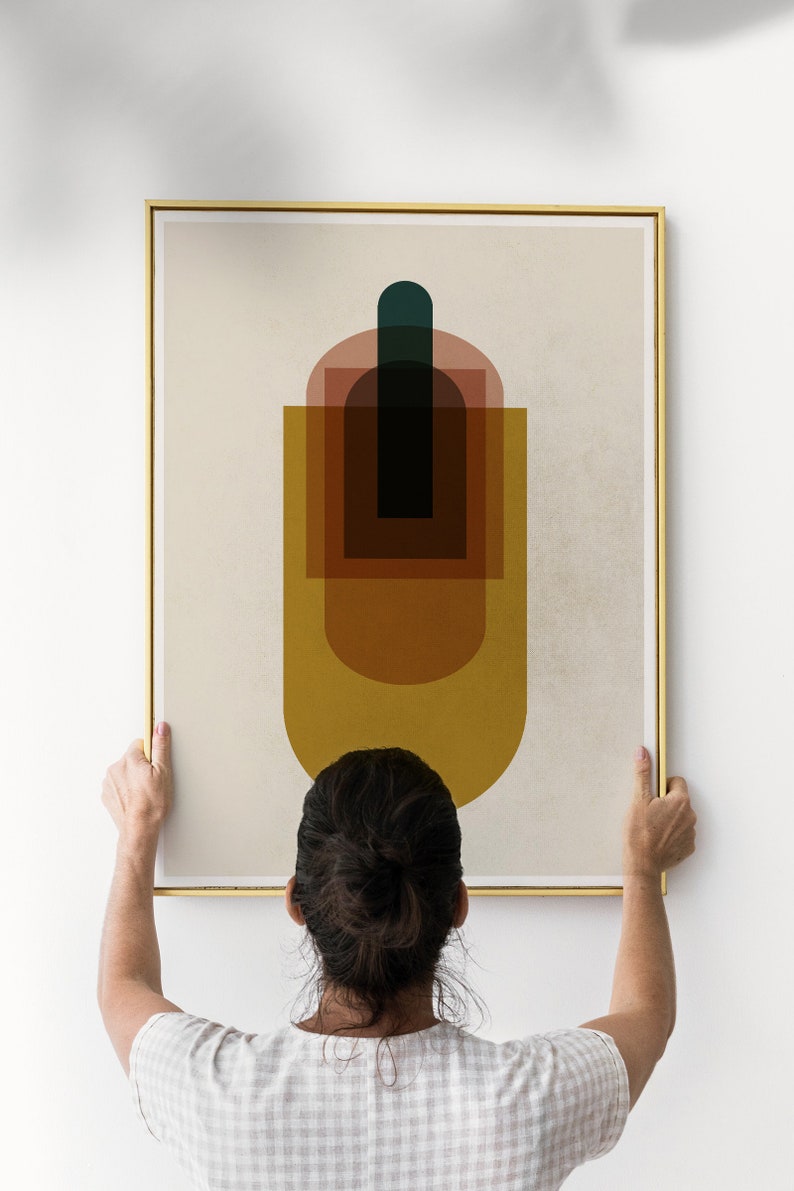 May include: A person hangs a framed abstract art print on a white wall. The print features a geometric design with overlapping shapes in shades of brown, yellow, red, and green.
