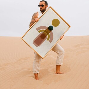 May include: A person holding a framed abstract print with geometric shapes in shades of pink, yellow, green, and brown. The print is on a white background and has a gold frame.