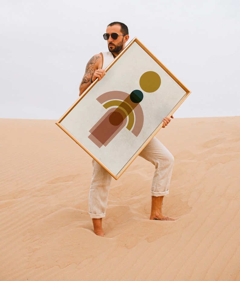 May include: A person holding a framed abstract print with geometric shapes in shades of pink, yellow, green, and brown. The print is on a white background and has a gold frame.