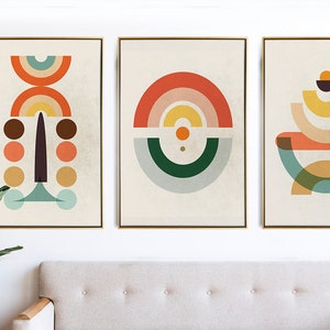 May include: Three framed abstract art prints with geometric shapes in orange, yellow, brown, and teal. The artwork is displayed above a beige sofa and next to a large green plant. The frames have a gold finish.
