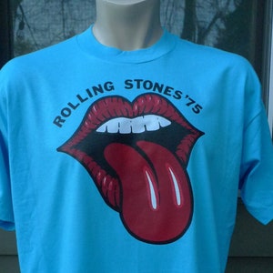 May include: A light blue t-shirt with a red and black Rolling Stones logo. The logo features a tongue sticking out of a mouth with the text "Rolling Stones '75".