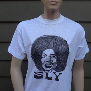May include: White t-shirt with a black and white illustration of Sly Stone, the lead singer of Sly and the Family Stone. The illustration shows Stone with an afro and a wide smile. The text "SLY" is printed below the illustration.