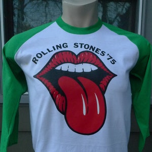 May include: White and green baseball style t-shirt with a red tongue and lips graphic and the text "Rolling Stones '75".