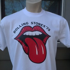 May include: White t-shirt featuring the Rolling Stones' iconic red lips and tongue logo from their 1975 tour. The graphic is in red, black, and white. The shirt is displayed on a mannequin.
