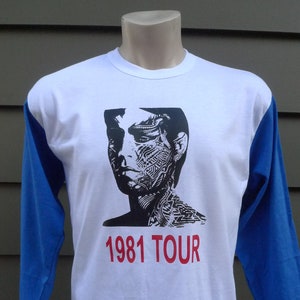 May include: A white and blue baseball t-shirt with a black and white image of a person's face and the text "1981 TOUR" in red.