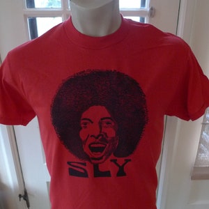 May include: Red t-shirt featuring a black graphic of a person with a large afro and the word "SLY" in black block letters. The shirt is displayed on a mannequin.
