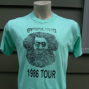 May include: A turquoise t-shirt with a black and white illustration of a man with a beard and sunglasses. The text "Grateful Dead" and "1986 Tour" is printed on the shirt.