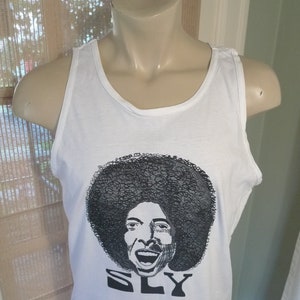 May include: White tank top with a black and white graphic of a person with an afro and the word "SLY" printed below. The tank top is displayed on a mannequin.