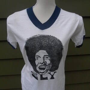 May include: White t-shirt with a navy blue v-neck collar and navy blue trim on the sleeves. The shirt features a black and white illustration of Sly Stone with the word "SLY" printed below.