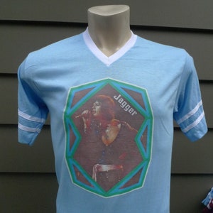 May include: Light blue t-shirt with white stripes on the sleeves. The shirt features a graphic of Mick Jagger in a geometric frame with the word "Jagger" printed on the frame.