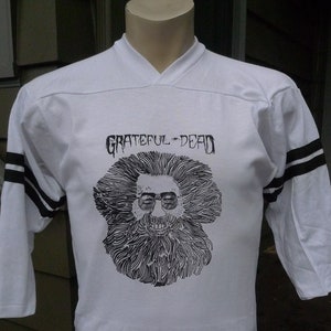 May include: White jersey-style shirt with black stripes on the sleeves. The shirt features a graphic of a bearded man wearing sunglasses and the text "Grateful Dead" in black. The shirt is displayed on a mannequin.