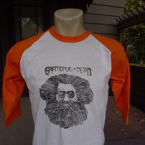 May include: White and orange baseball tee shirt with a black and white illustration of a man with a beard and the text "Grateful Dead" printed on the front.