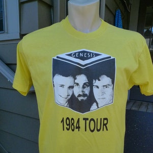 May include: Yellow t-shirt with a black and white image of the band Genesis. The image includes the band's name and the text "1984 Tour".