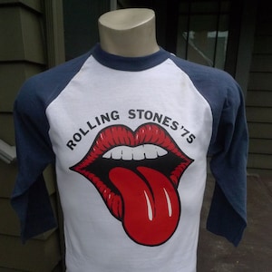 May include: White baseball tee shirt with blue sleeves. The shirt has a red and white graphic of the Rolling Stones tongue logo with the text "Rolling Stones '75".