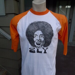 May include: White t-shirt with orange sleeves and a black and white illustration of Sly Stone's face with the word "SLY" printed below.