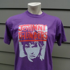 Może przedstawiać: Fioletowy T-shirt z białym obrazkiem Johnny'ego Thundersa i napisem "JOHNNY THUNDERS" w kolorze różowym.