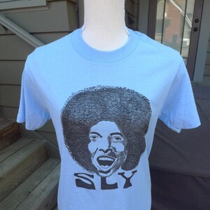 May include: A light blue t-shirt with a black and white illustration of Sly Stone, a singer, with the word "SLY" printed below the illustration.