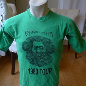 May include: Green t-shirt with a black and white graphic of Jerry Garcia from the Grateful Dead. The text "Grateful Dead" and "1980 Tour" is printed on the shirt.