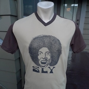May include: A beige t-shirt with brown sleeves features a black and white illustration of a person with an afro and the word "SLY" printed below.