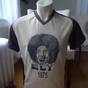 May include: A beige and brown short-sleeved t-shirt with a black and white image of Sly Stone and the text "SLY 1975".