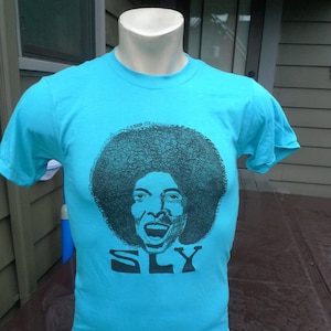 May include: A turquoise blue t-shirt with a black and white graphic of a person with an afro and the word "SLY" printed below.