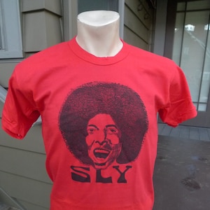 May include: Red t-shirt with a black and white illustration of Sly Stone with an afro and the word "SLY" printed below.
