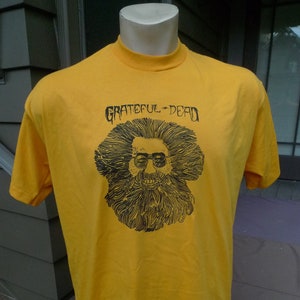 May include: A yellow t-shirt with a black and white illustration of Jerry Garcia, the lead singer of the Grateful Dead, and the text "Grateful Dead".
