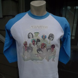 May include: A white baseball tee shirt with blue sleeves. The shirt features a graphic of the Rolling Stones band members with insect heads. The text "METAMORPHOSIS" and "ROLLING STONES" are printed on the shirt.