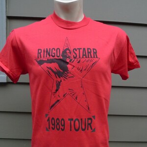 Może przedstawiać: Czerwony T-shirt z czarną gwiazdą i nadrukiem "RINGO STARR 1989 TOUR" z przodu.
