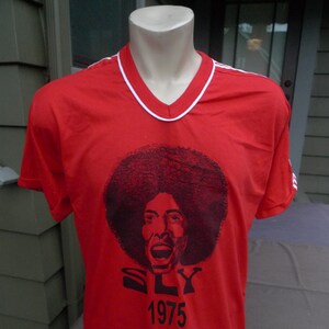 May include: Red t-shirt with a black and white image of a person with an afro and the text "SLY 1975".