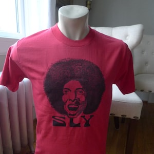 May include: Red t-shirt with a black and white image of Sly Stone with an afro and the word "SLY" printed below.