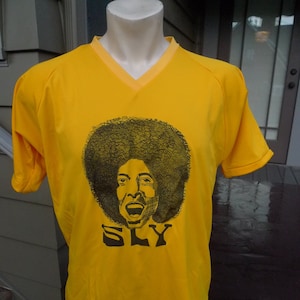 May include: A yellow t-shirt with a black and white image of Sly Stone, the lead singer of Sly and the Family Stone. The shirt has a v-neck and the image is centered on the front of the shirt.