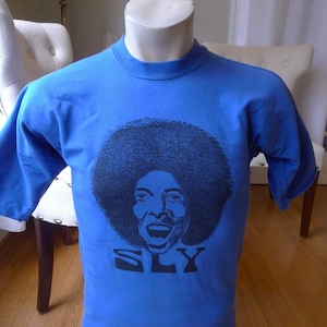 May include: A blue t-shirt with a black and white image of Sly Stone, the lead singer of Sly and the Family Stone. The shirt has the word "SLY" printed in black below the image.