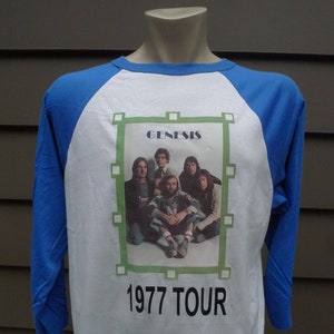 May include: White and blue baseball style t-shirt with a picture of the band Genesis from their 1977 tour. The band members are pictured in a group shot with the text "Genesis" and "1977 Tour" printed on the shirt.