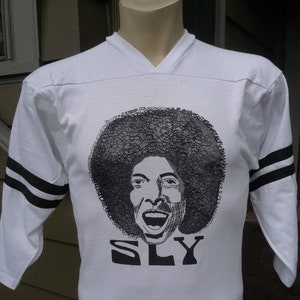 May include: White long-sleeve jersey with black stripes on the sleeves. The jersey features a black and white illustration of a person with an afro and the word "SLY" printed below the illustration.