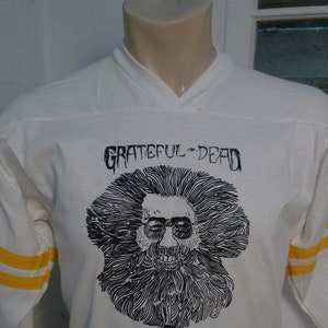 May include: White long-sleeve t-shirt with a black and white illustration of a man with a beard and sunglasses. The text "GRATEFUL DEAD" and "1979 TOUR" is printed on the shirt. The shirt has yellow and white striped sleeves.