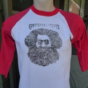 May include: A white and red baseball tee shirt with a black and white illustration of a man with a beard and sunglasses. The text "Grateful Dead" is printed on the shirt.