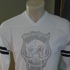 Może przedstawiać: Biała koszulka z nadrukiem srebrnego brokatu przedstawiającym czaszkę z rogami i tekstem "Motorhead 1981 Tour".