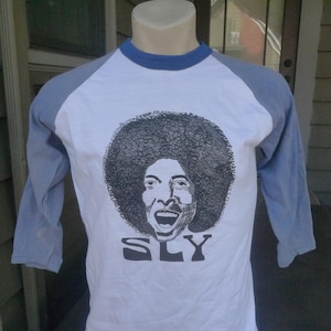 May include: A white and blue baseball tee shirt with a black and white illustration of Sly Stone's face and the word "SLY" printed on the front.
