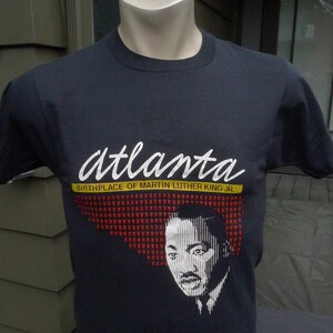 Puede incluir: Una camiseta negra con un gráfico blanco de Martin Luther King Jr. y el texto "Atlanta, Birthplace of Martin Luther King Jr."