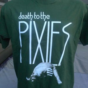 Può includere: T-shirt verde scuro con la scritta bianca "death to the PIXIES" e una grafica bianca. La maglietta è esposta su un manichino.