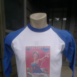 May include: A white and blue baseball style t-shirt with a vintage Rolling Stones Tour graphic featuring a woman in a blue jumpsuit and a red background. The text "Rolling Stones Tour" is printed at the top of the graphic.